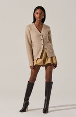 A woman is wearing a beige, chunky knit cardigan with large front pockets and wooden buttons. She has on a beige, voluminous skirt and tall, black knee-high boots with high heels.