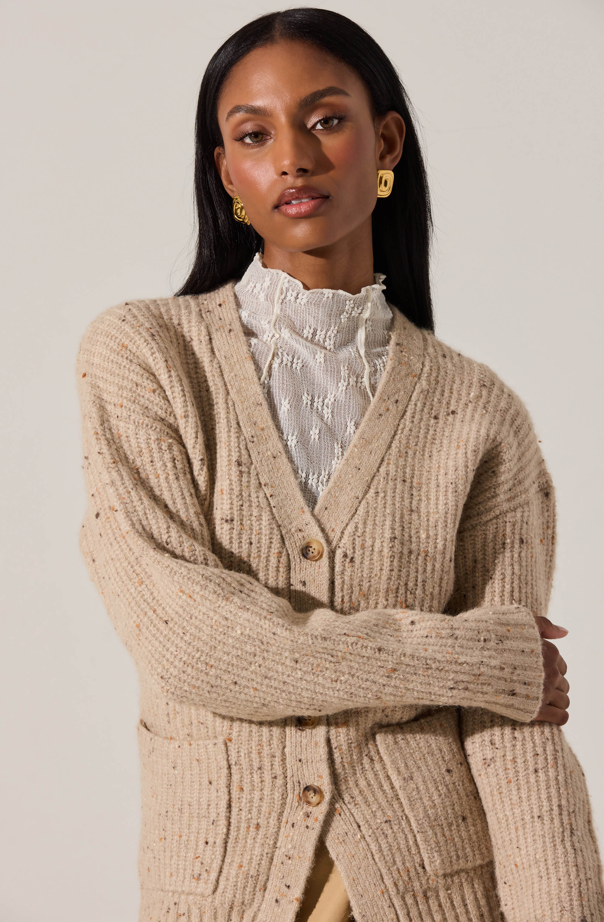 A woman is wearing a beige, chunky knit cardigan with large buttons down the front. Underneath, she has on a white, high-neck blouse with ruffled detailing at the collar. She accessorizes with gold, geometric-shaped earrings.