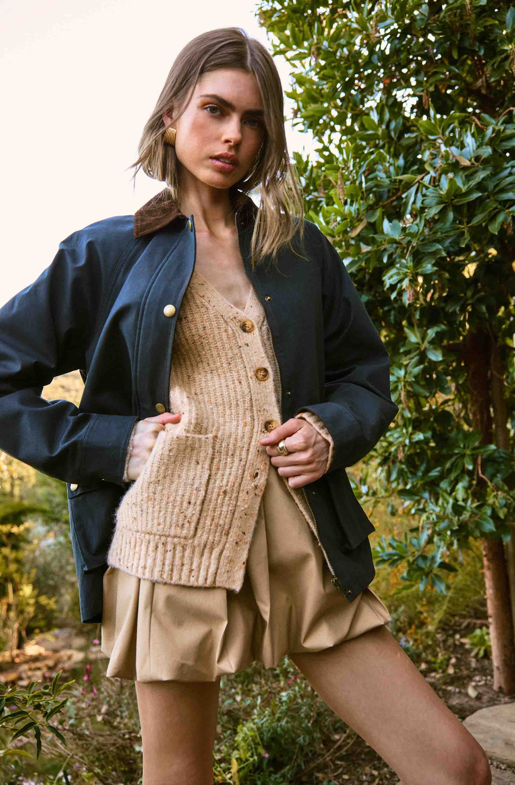 A young woman is wearing a beige, chunky knit cardigan with large buttons over a light-colored top, paired with beige high-waisted shorts that have a puffy, pleated design. She has a dark navy jacket with a brown collar draped over her shoulders. The background features green foliage, indicating an outdoor setting.