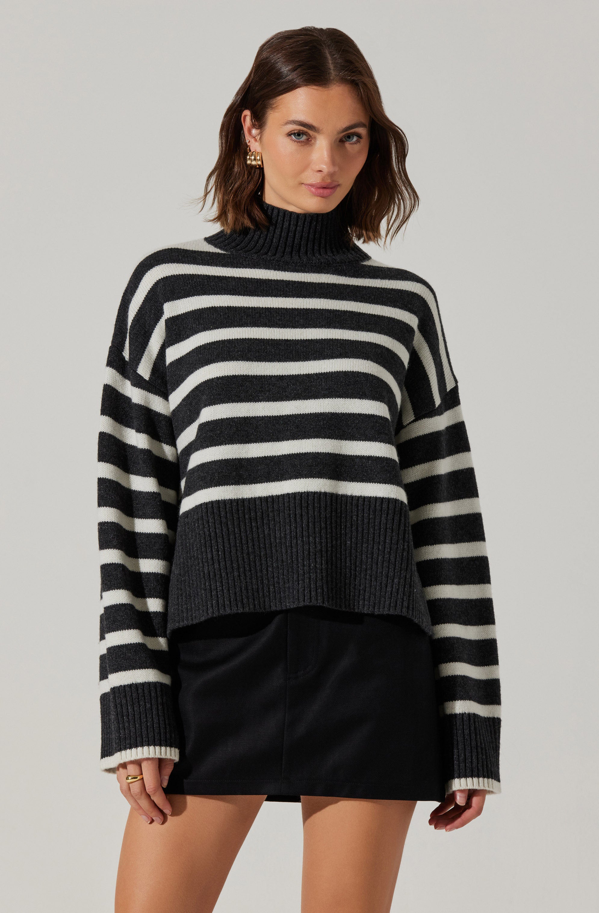 A woman is wearing a black and white horizontally striped turtleneck sweater with a chunky knit design. The sweater features wider black and white stripes and a ribbed hem and cuffs. She pairs the sweater with a black leather mini skirt.