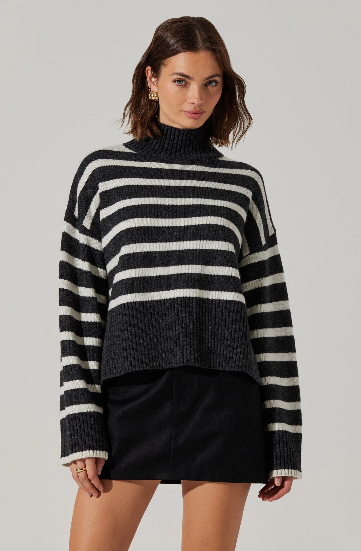 A woman is wearing a black and white horizontally striped turtleneck sweater with a chunky knit design. The sweater features wider black and white stripes and a ribbed hem and cuffs. She pairs the sweater with a black leather mini skirt.