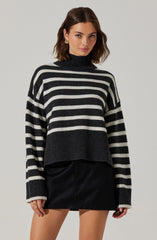 A woman is wearing a black and white horizontally striped turtleneck sweater with a chunky knit design. The sweater features wider black and white stripes and a ribbed hem and cuffs. She pairs the sweater with a black leather mini skirt.