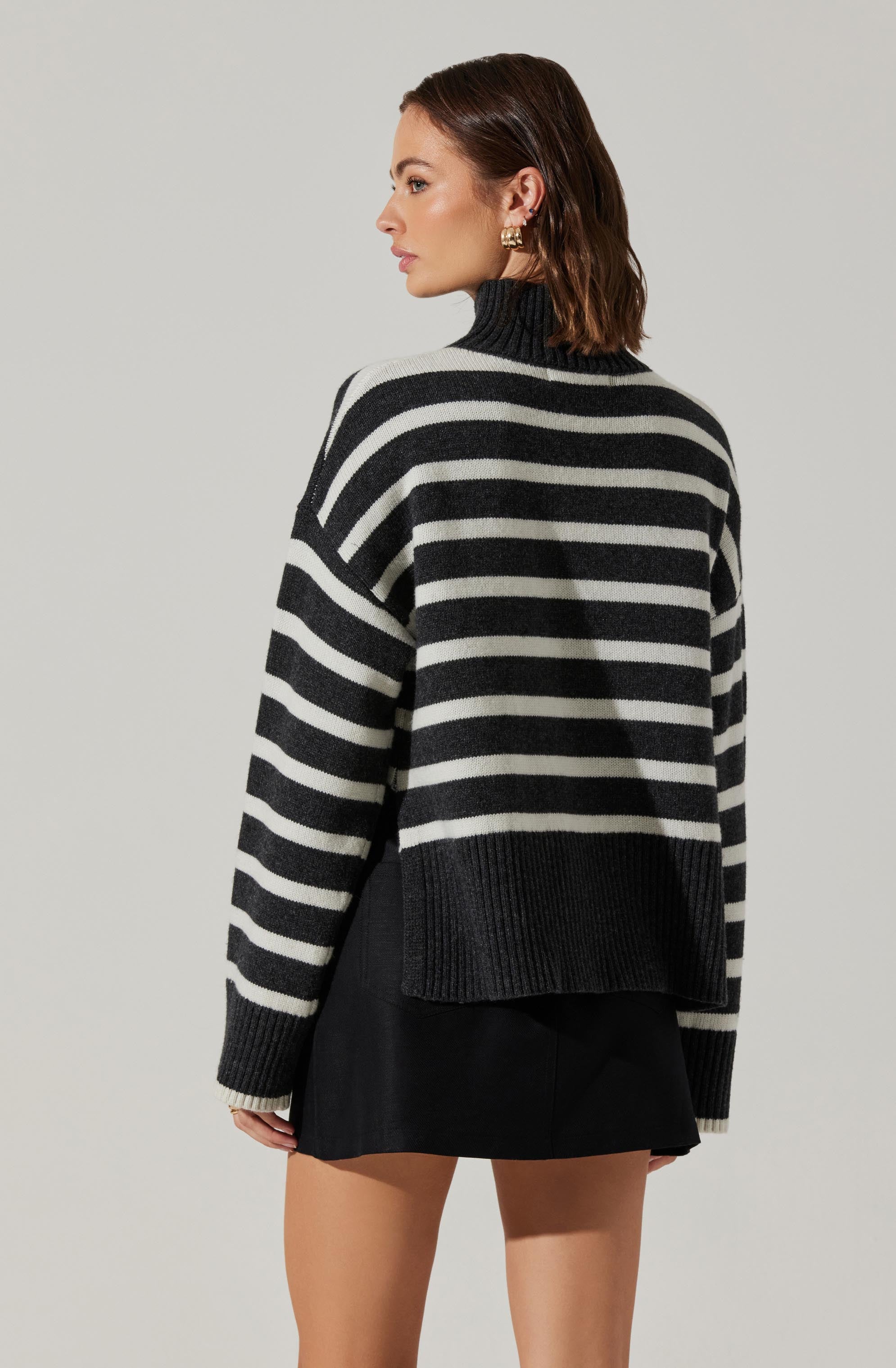 A woman is wearing a loose-fitting, long-sleeved sweater with wide horizontal black and white stripes, a high turtleneck collar, and ribbed cuffs and hem. She pairs it with a black skirt.