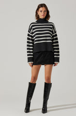 A woman is wearing a loose-fitting, oversized sweater with horizontal black and gray stripes, a high ribbed neckline, and ribbed cuffs. She pairs the sweater with a short black skirt and tall black leather boots that reach just below her knees.