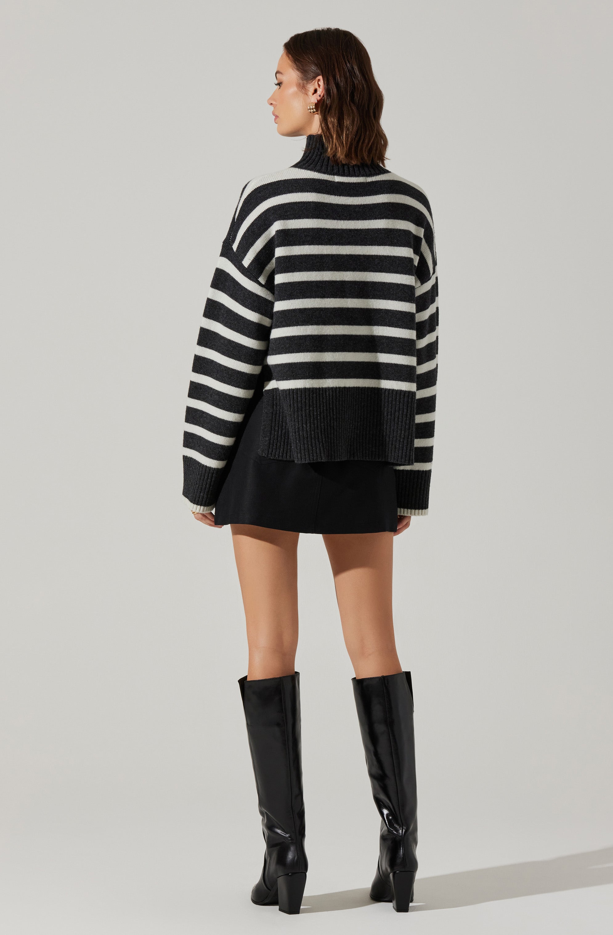 A woman is wearing an oversized black and white horizontally striped sweater with a high, ribbed neckline. She is also wearing a short black skirt and knee-high black glossy leather boots with chunky heels.