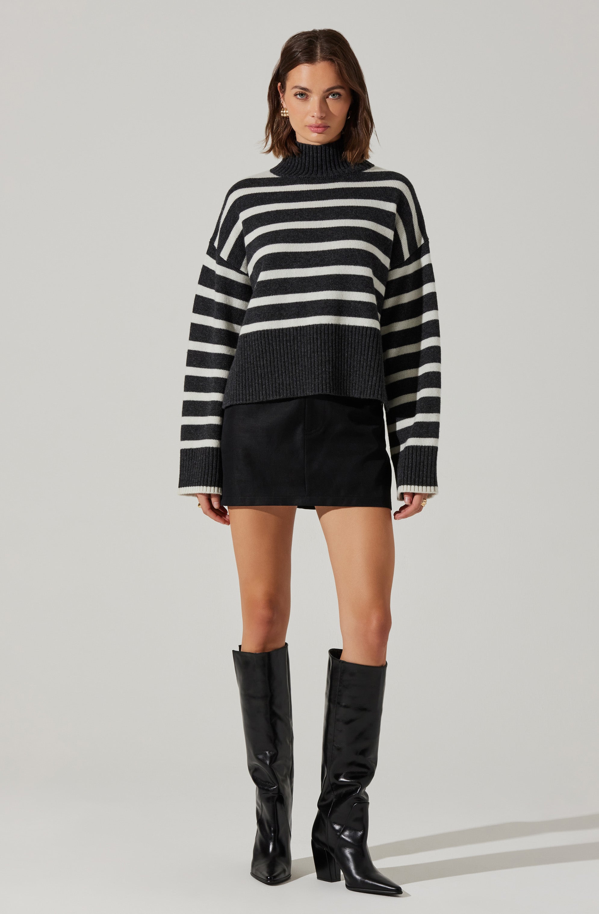 A woman is wearing a black and gray striped turtleneck sweater with oversized sleeves, paired with a black mini skirt and knee-high black shiny leather boots with pointed toes and chunky heels.