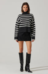 A woman is wearing a black and gray striped turtleneck sweater with oversized sleeves, paired with a black mini skirt and knee-high black shiny leather boots with pointed toes and chunky heels.