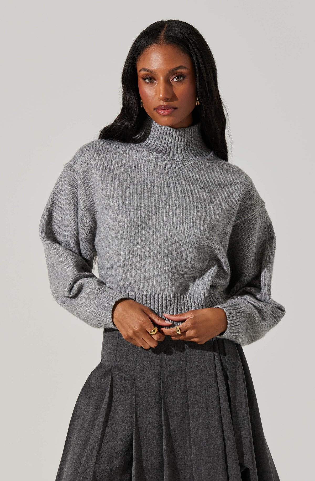 A woman is wearing a chunky, light gray knit sweater with a high turtleneck and long, slightly puffed sleeves. She pairs it with a high-waisted, dark gray pleated skirt.