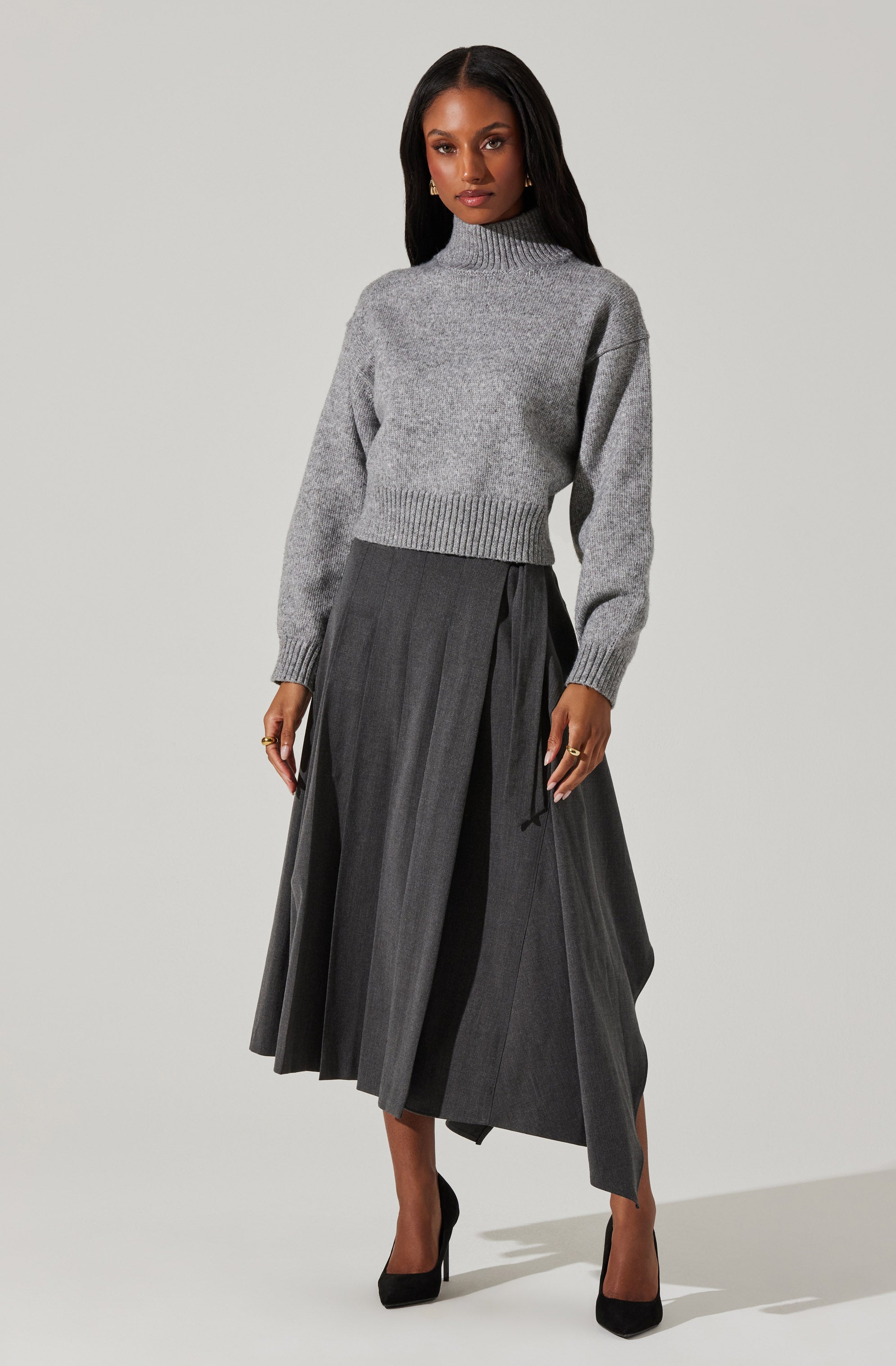 A woman is wearing a light gray, high-neck knit sweater with long sleeves and ribbed cuffs. She pairs it with a dark gray, pleated, ankle-length skirt that has a wrap-style design and a tie in the front. She is also wearing black high-heeled shoes.