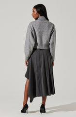 A woman is wearing a grey knit sweater with a high turtleneck and long, slightly puffed sleeves. She pairs it with a high-waisted, dark grey pleated midi skirt that has an asymmetrical hemline. She completes the look with black high-heeled shoes.