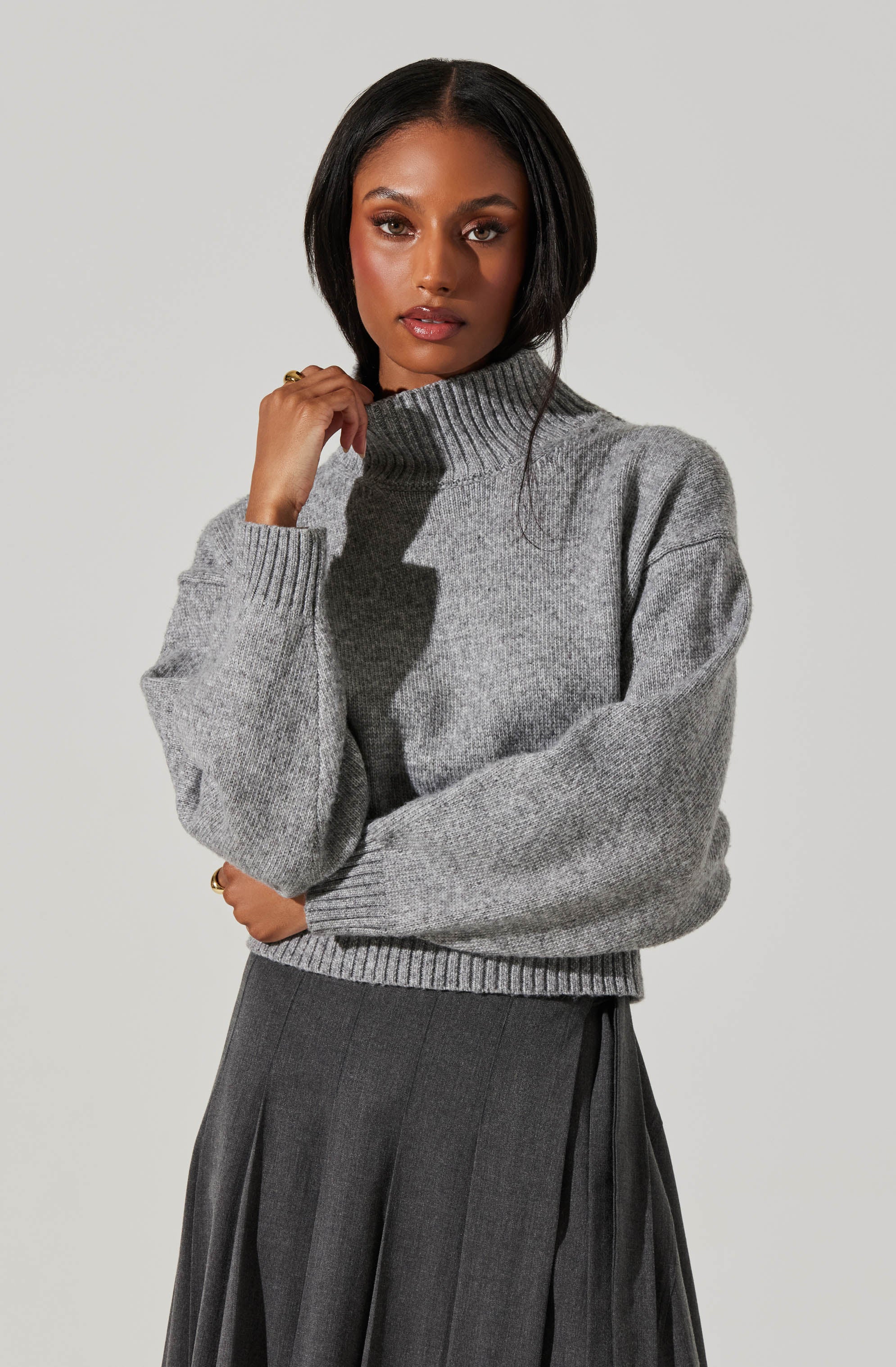 A woman is wearing a gray turtleneck sweater with a ribbed collar, cuffs, and hem. The sweater appears to be made of a soft, knit material and has a slightly oversized fit. She pairs it with a dark gray, pleated skirt that has a high waist and a subtle textured pattern.