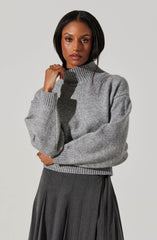 A woman is wearing a gray turtleneck sweater with a ribbed collar, cuffs, and hem. The sweater appears to be made of a soft, knit material and has a slightly oversized fit. She pairs it with a dark gray, pleated skirt that has a high waist and a subtle textured pattern.