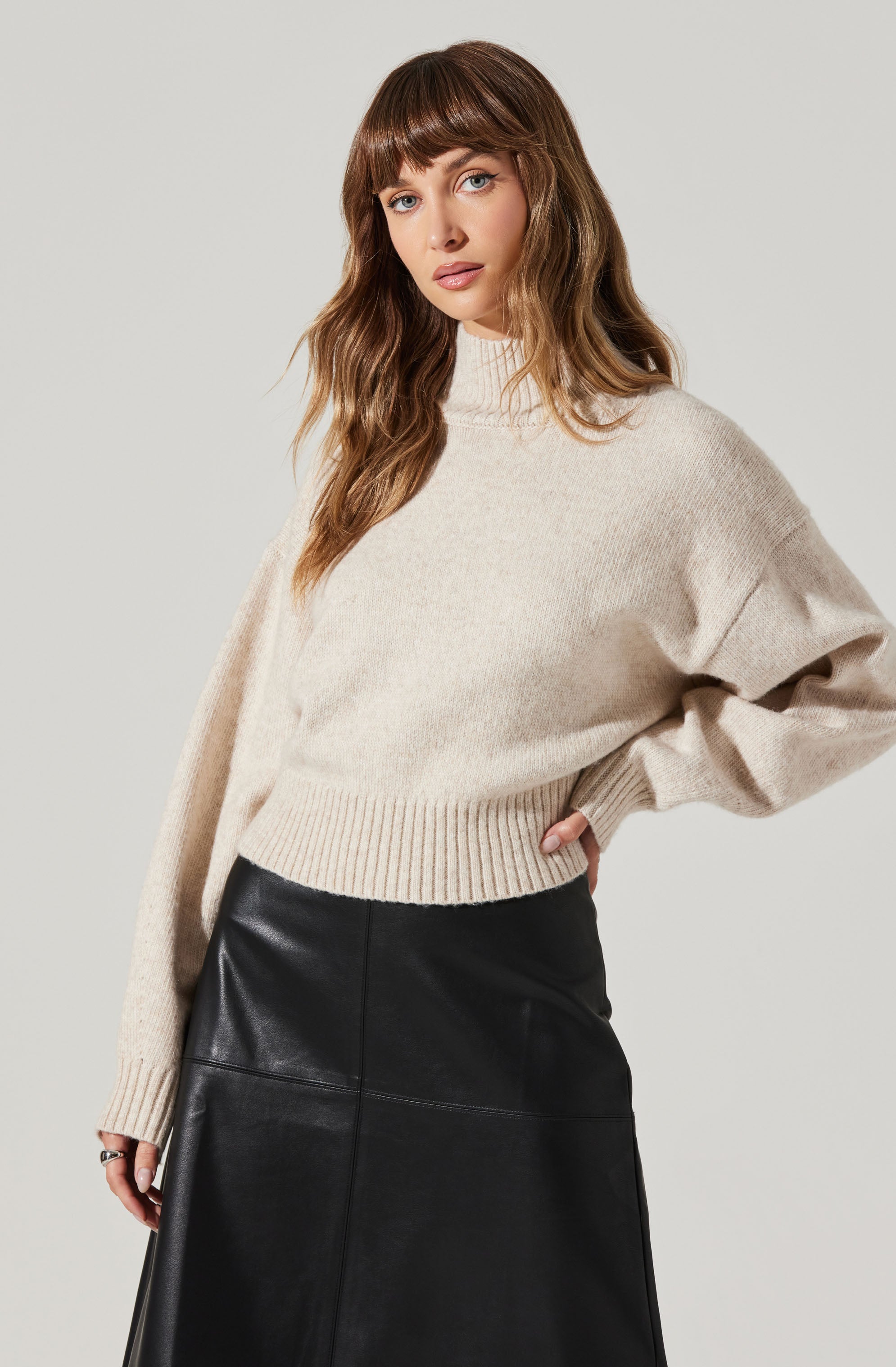 A woman is wearing an oversized, cream-colored knit sweater with a high neckline and long sleeves. She has paired it with a black leather skirt that has a slightly flared silhouette.