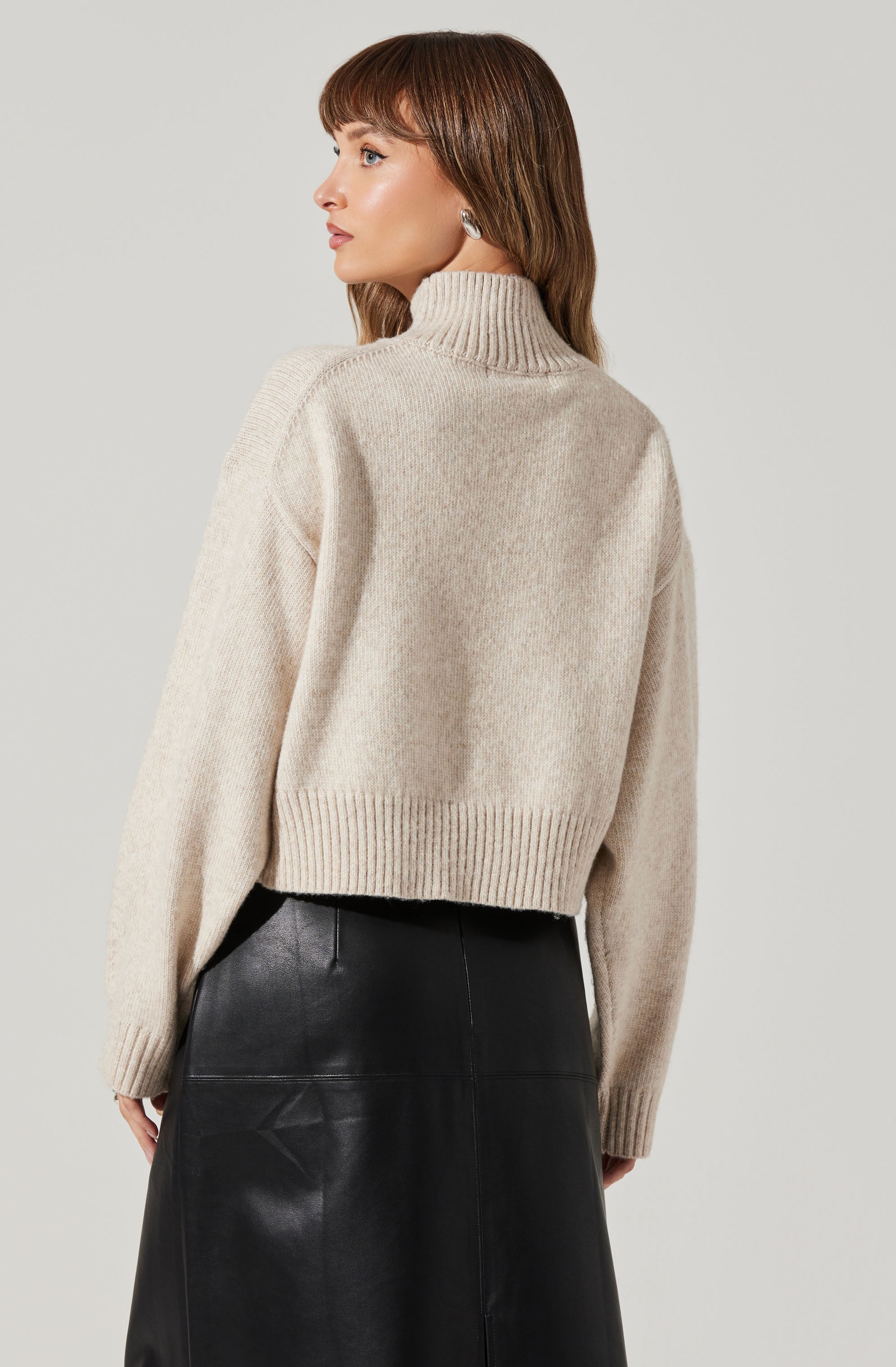 A woman is wearing a beige, high-neck, loose-fitting knit sweater with ribbed cuffs and hem. She is also dressed in a high-waisted black leather skirt.