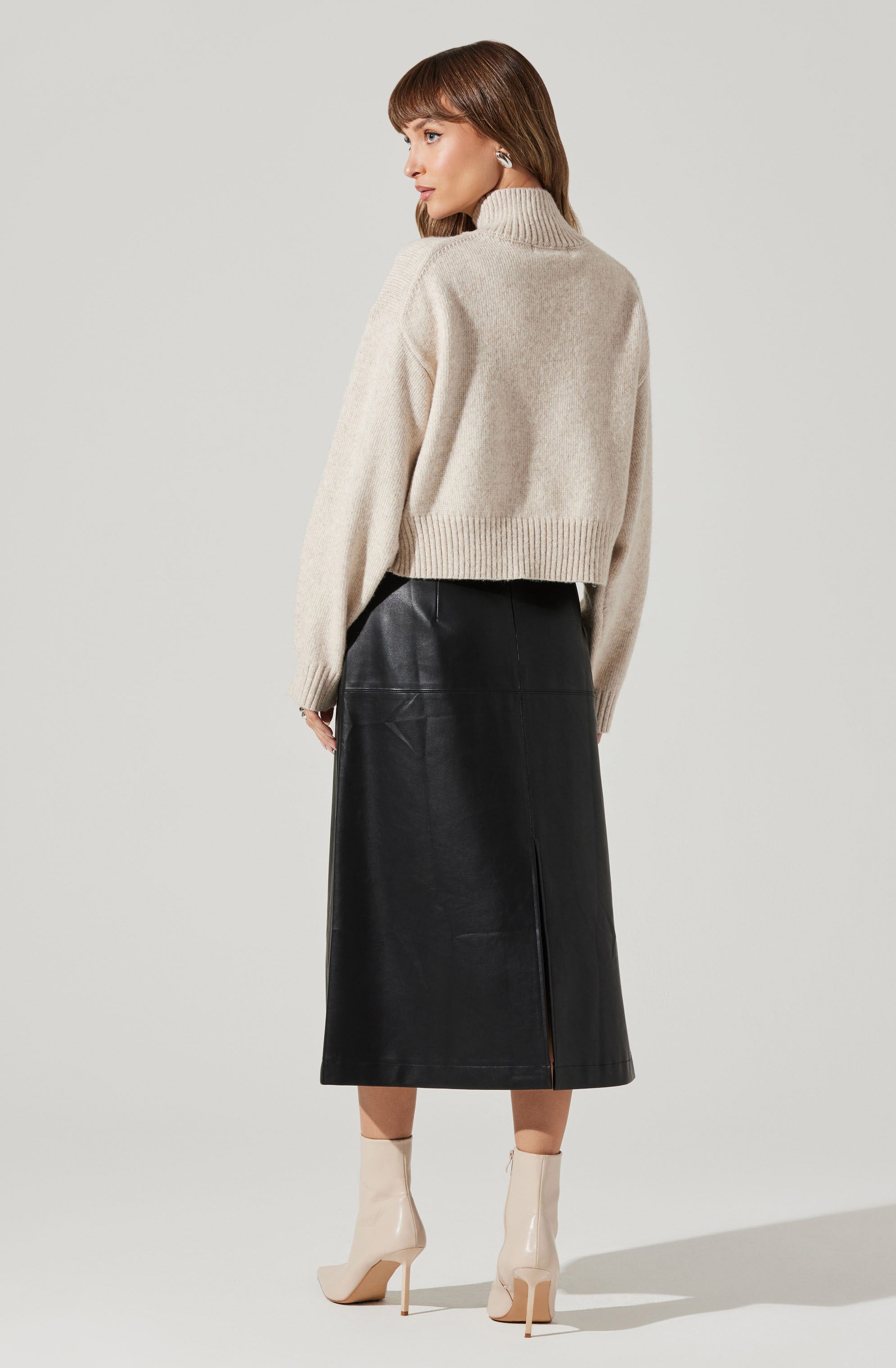 A woman is wearing a chunky, oversized, beige turtleneck sweater with ribbed cuffs and hem. She pairs it with a high-waisted, black leather midi skirt that has a front slit. She completes the outfit with shiny, beige, heeled ankle boots.