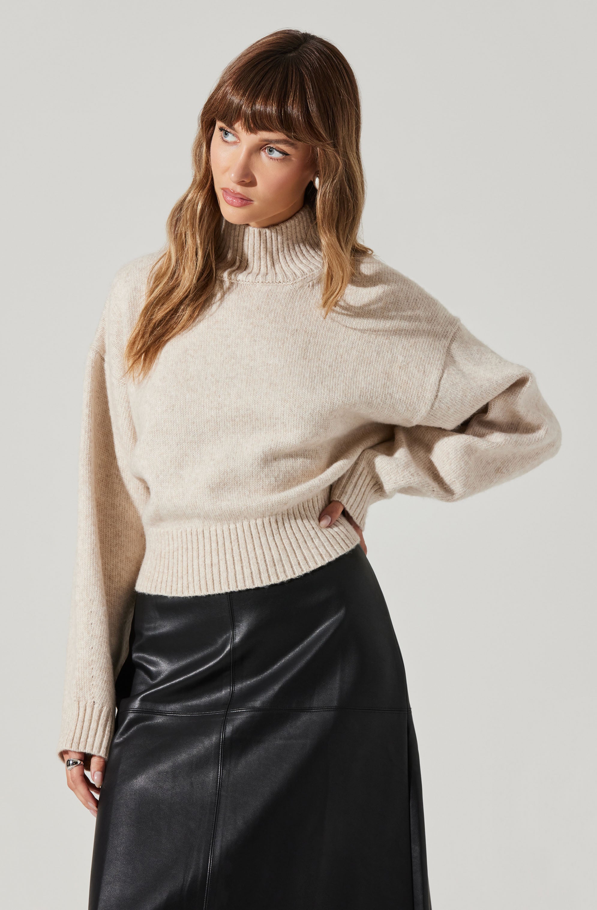 A woman is wearing a beige, oversized, high-neck knitted sweater with ribbed cuffs and hem, paired with a long black leather skirt.