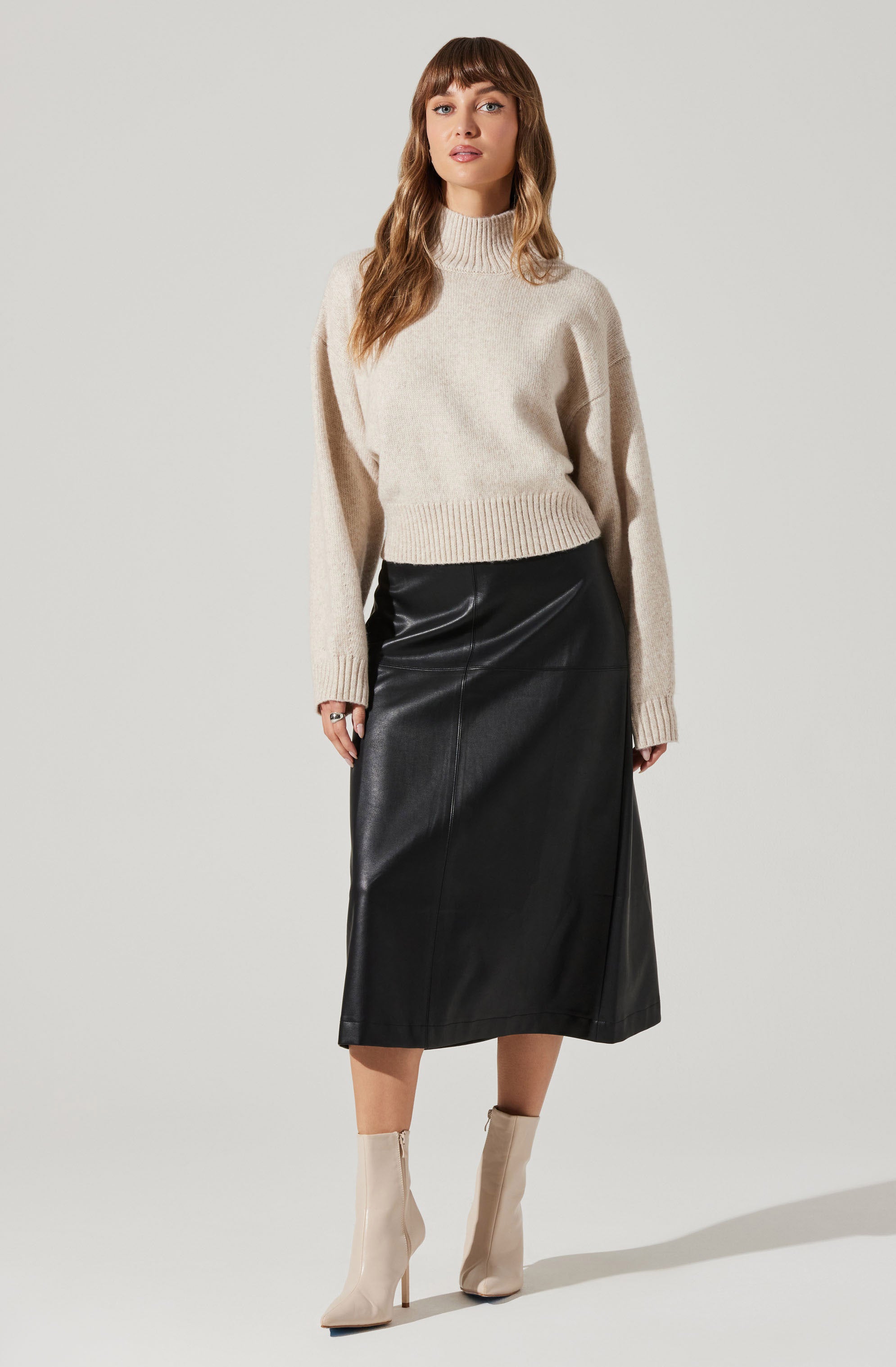 A woman is wearing a beige, high-neck, knitted sweater with ribbed cuffs and hem, paired with a long black leather skirt that falls below the knees. She has on light-colored ankle boots with high heels.