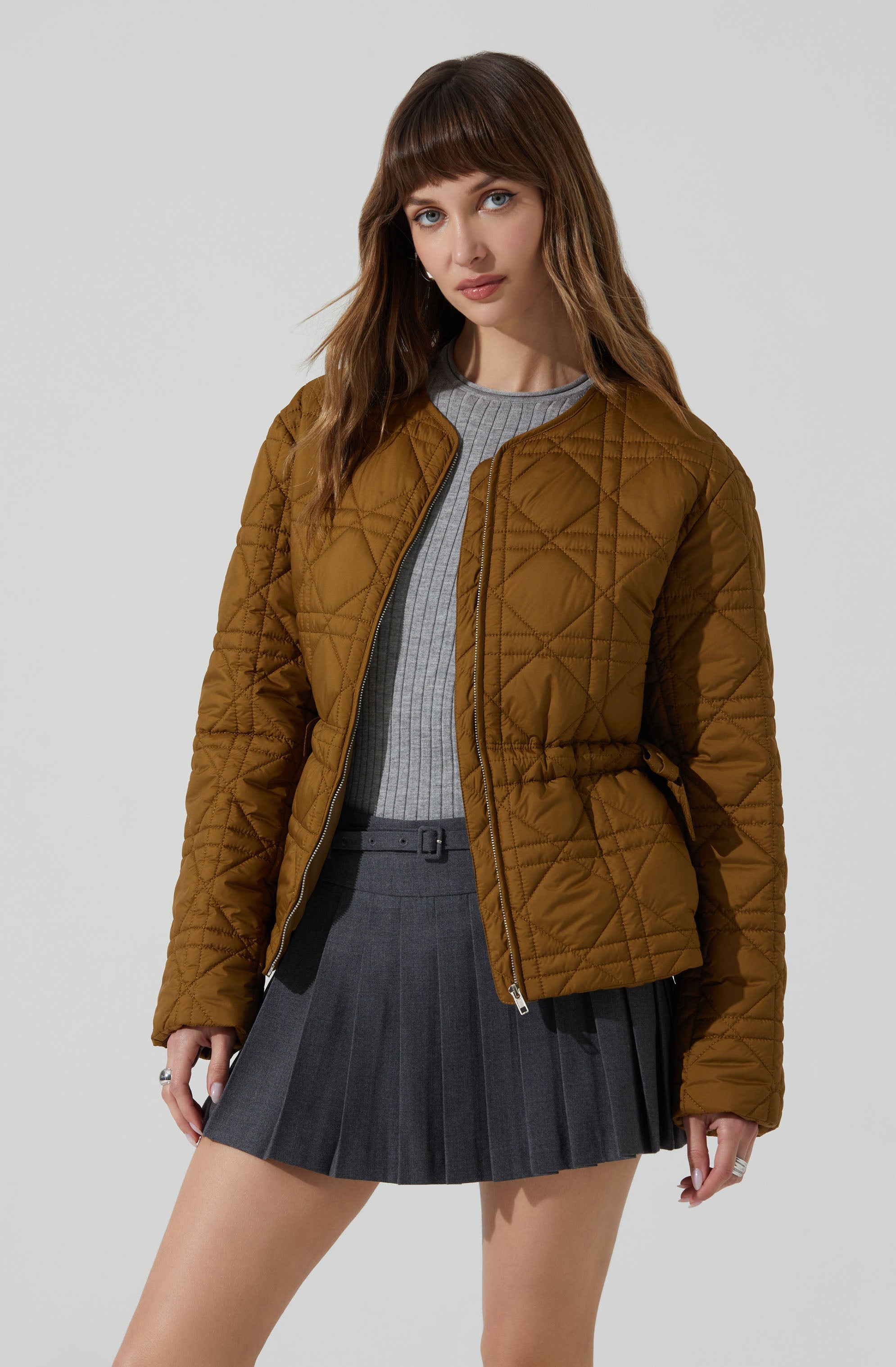 A woman is wearing a brown, quilted, zip-up jacket with a drawstring waist, paired with a light gray ribbed sweater and a dark gray pleated skirt.