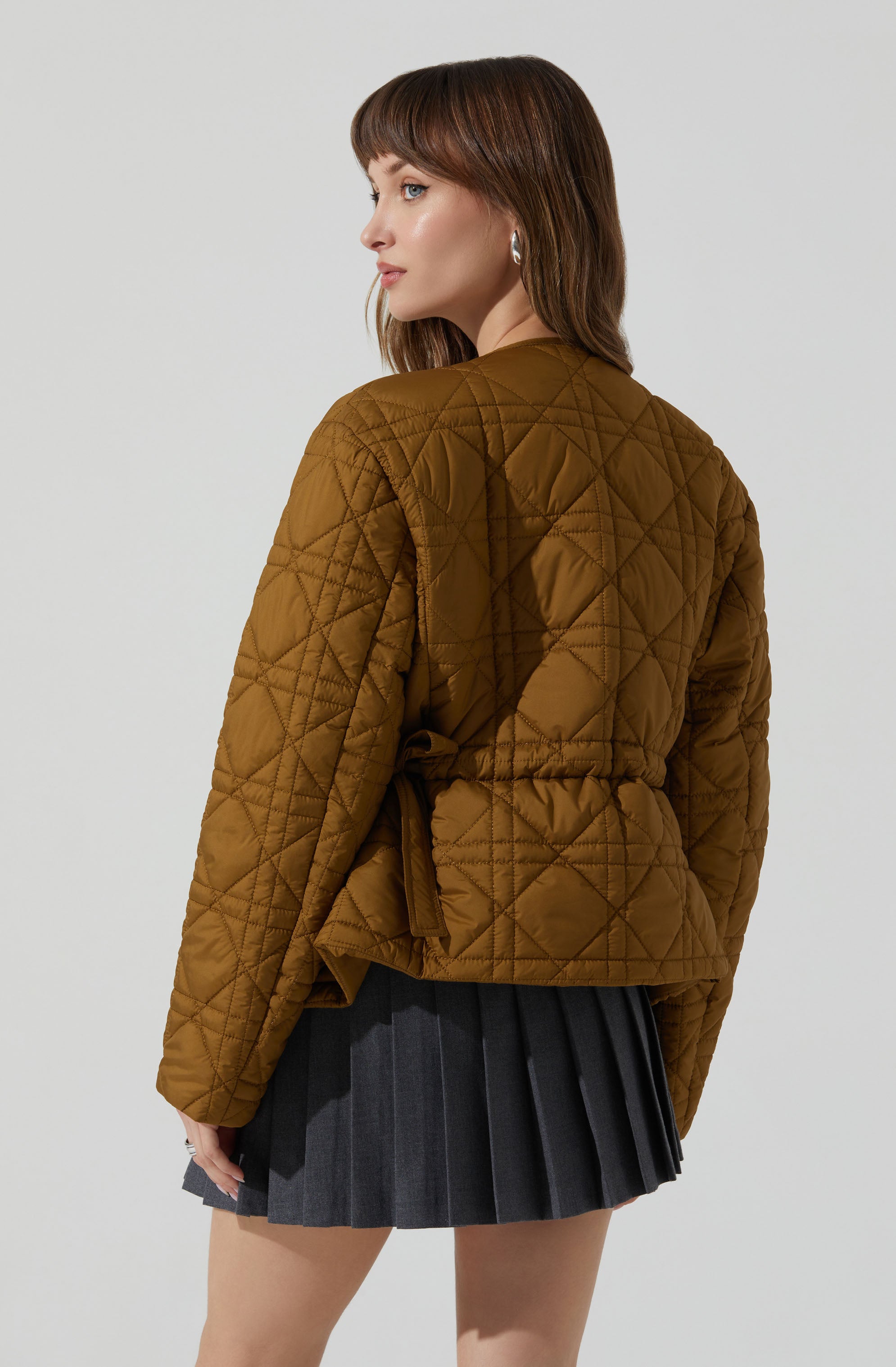 A woman is wearing a brown quilted jacket with diamond and diagonal stitch patterns, featuring a belted waist. She pairs it with a dark gray pleated skirt.