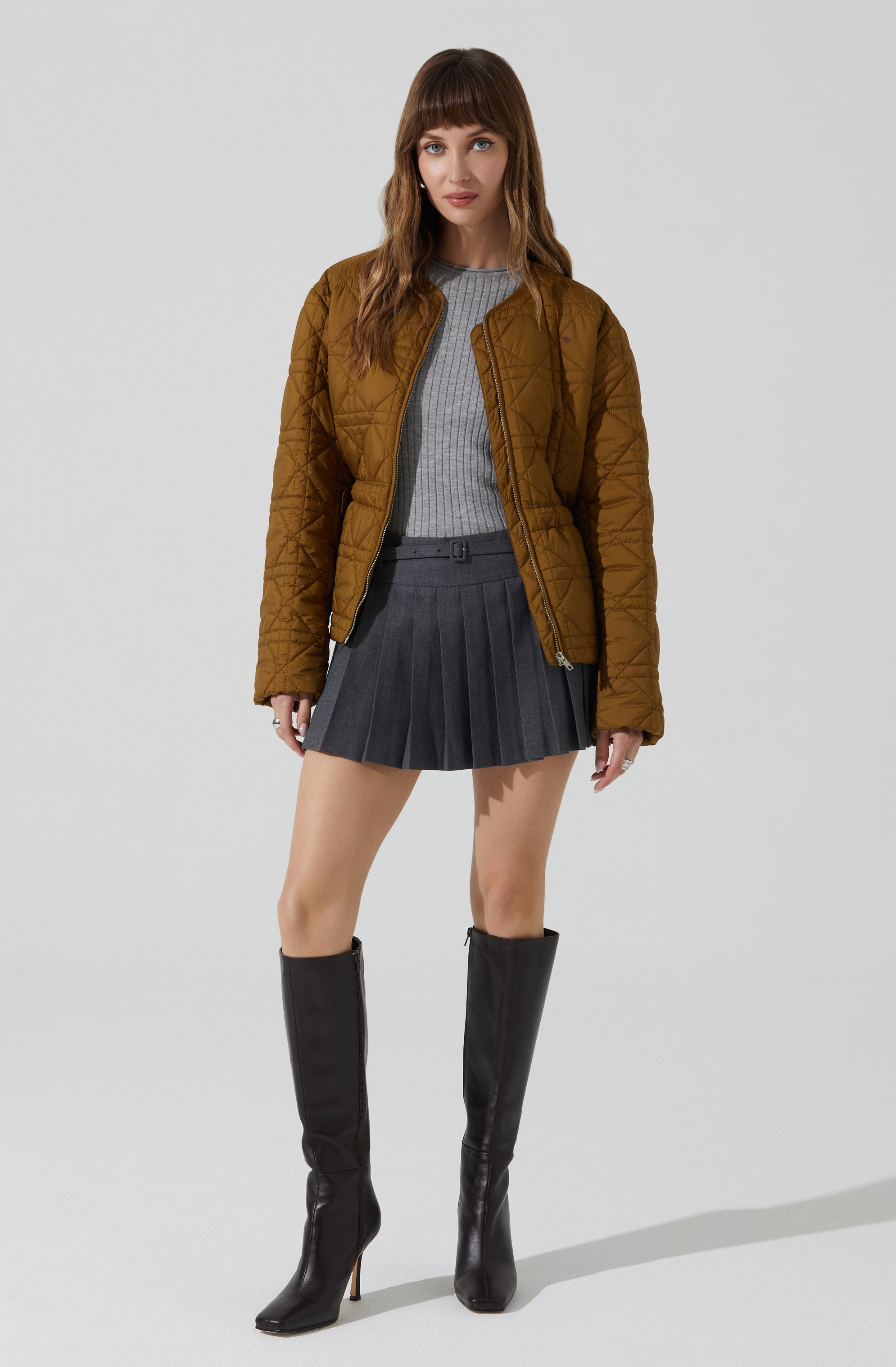 A woman is wearing a brown quilted jacket with a front zipper, a gray ribbed sweater, a dark gray pleated skirt, and black knee-high boots with high heels.