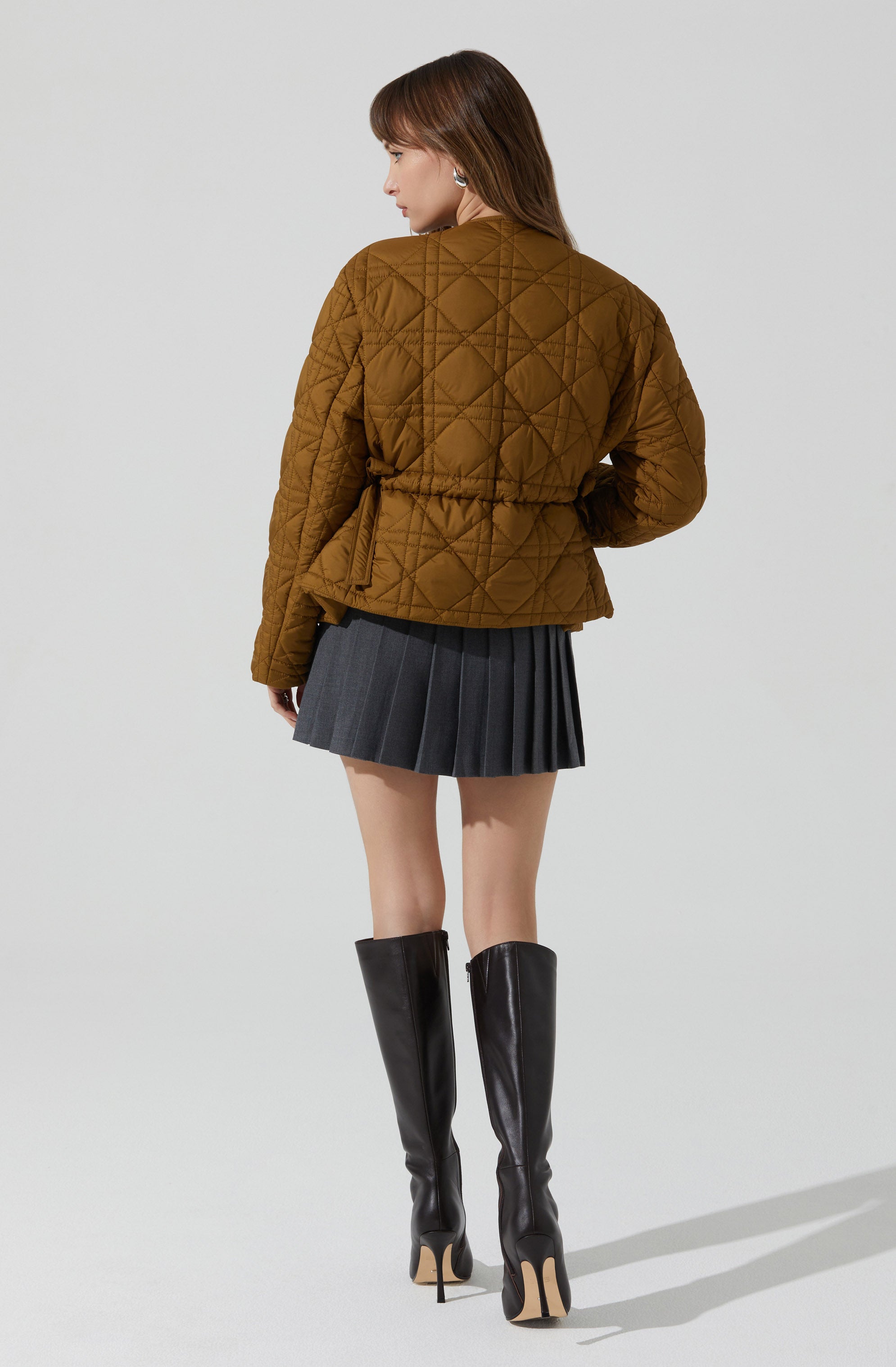 A woman is wearing a brown quilted jacket with diamond and diagonal stitching patterns. She has paired it with a dark gray pleated mini skirt and tall black leather high-heeled boots that reach just below her knees.