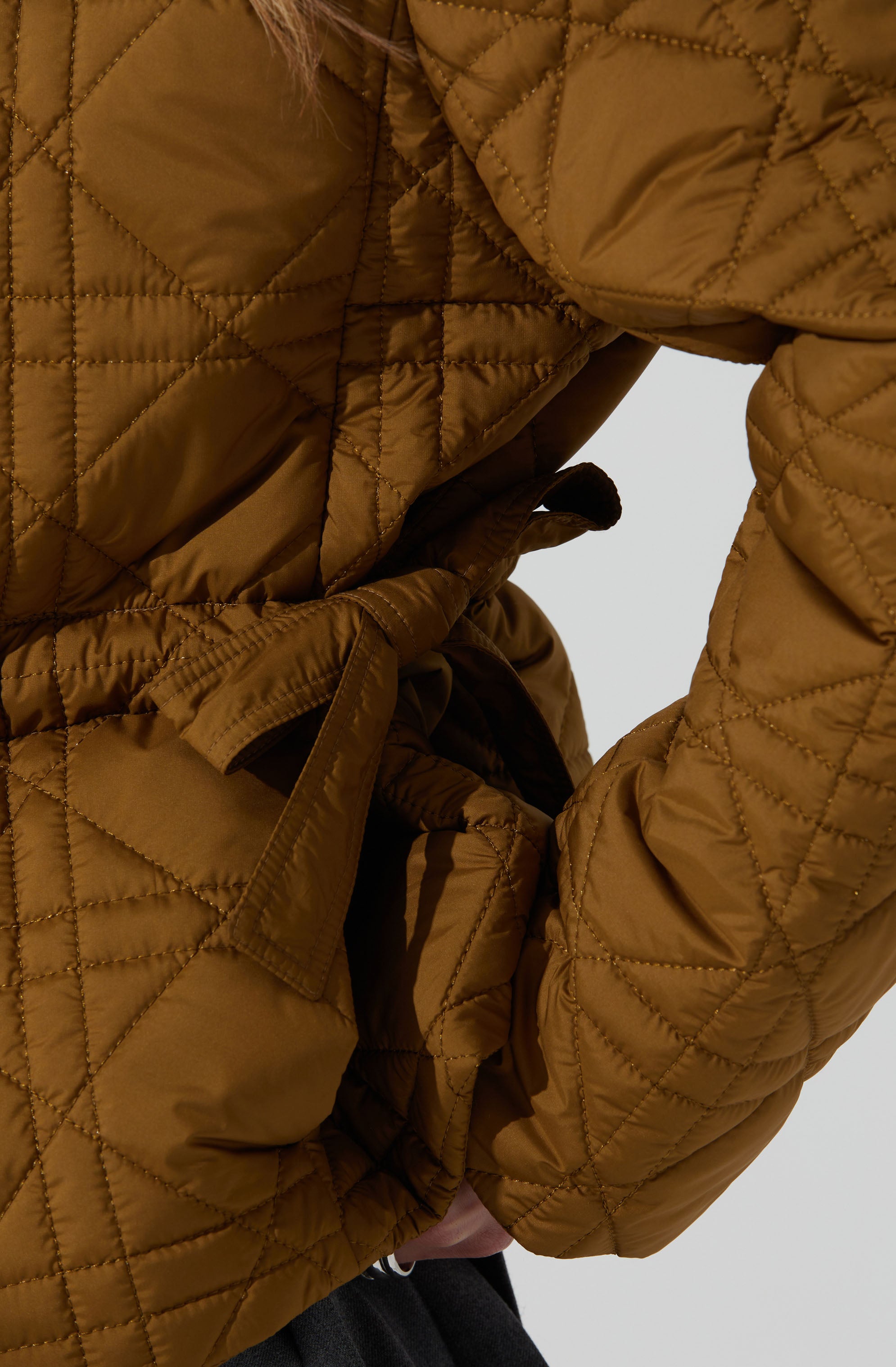 A person is wearing a quilted, tan-colored jacket with a diamond stitch pattern. The jacket features a fabric belt tied in a bow at the waist and has puffy, insulated sleeves. The overall style appears warm and padded, suitable for cold weather.