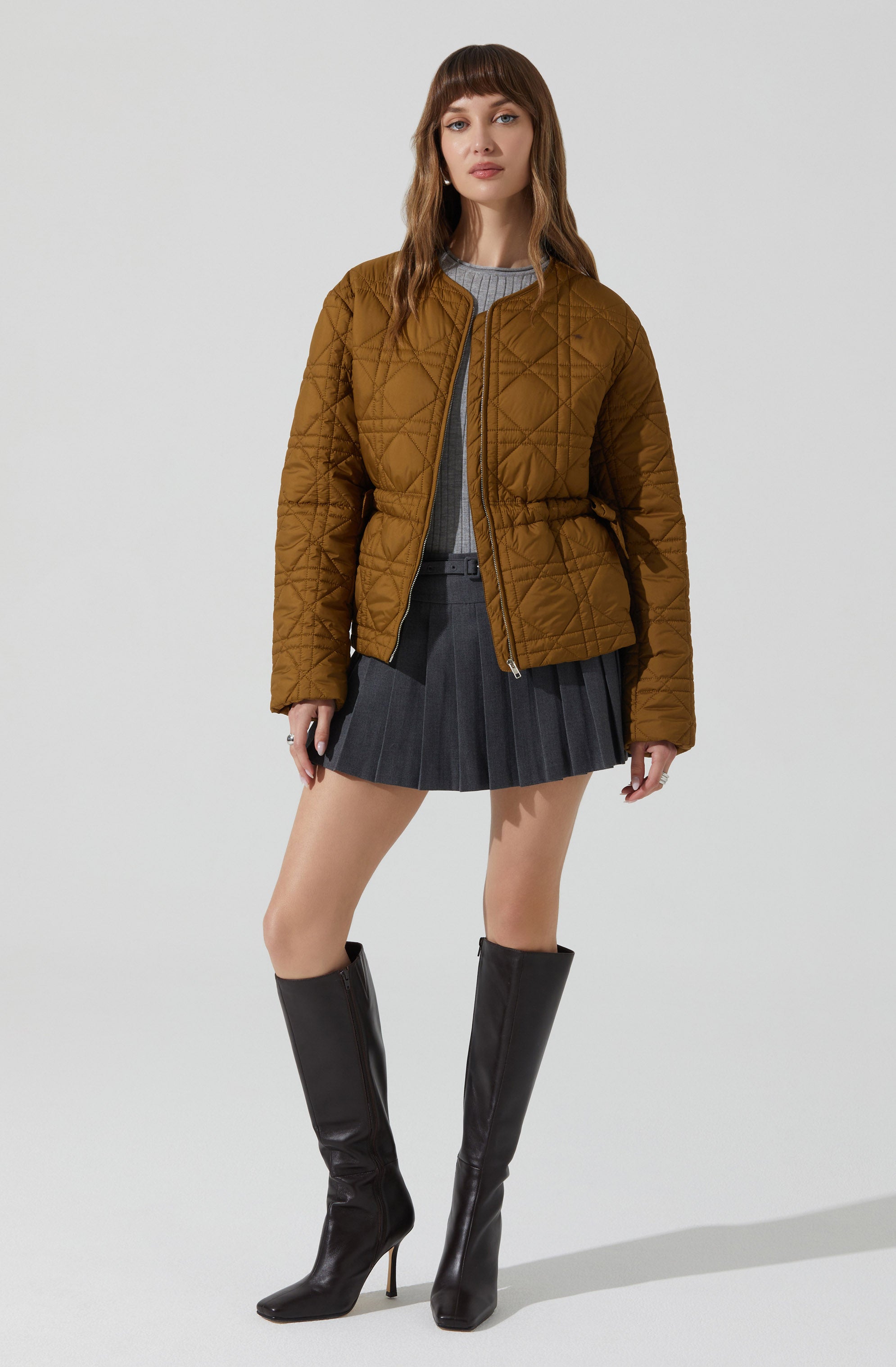 A woman is wearing a brown quilted jacket with a front zipper, a gray top underneath, a dark gray pleated skirt, and black knee-high pointed boots with high heels.