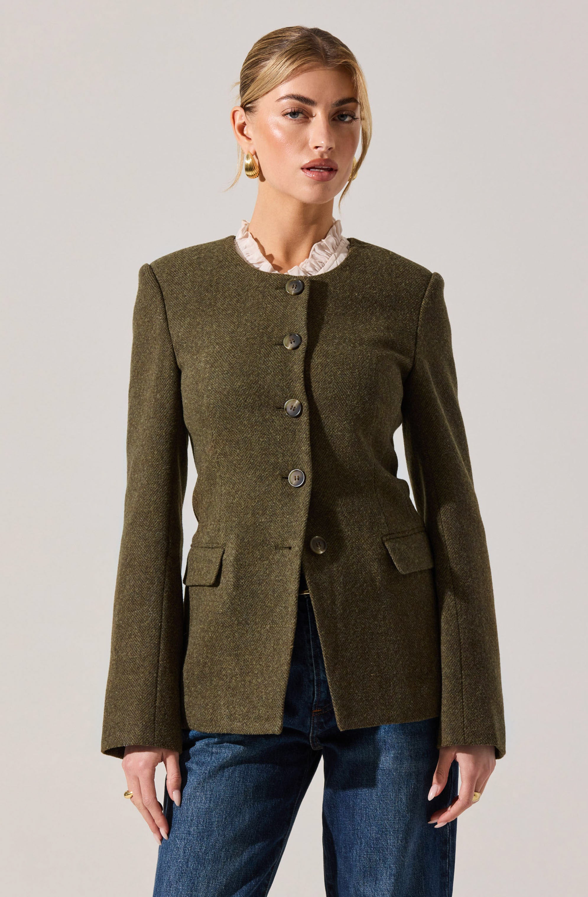A woman is wearing an olive green tailored blazer with a round neckline, featuring four large buttons down the front and two small flap pockets at the waist. She has a ruffled light beige or cream-colored blouse underneath the blazer, which has a high ruffled collar visible above the blazer's neckline. She is also wearing medium-wash blue jeans.
