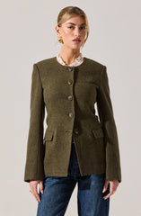 A woman is wearing an olive green tailored blazer with a round neckline, featuring four large buttons down the front and two small flap pockets at the waist. She has a ruffled light beige or cream-colored blouse underneath the blazer, which has a high ruffled collar visible above the blazer's neckline. She is also wearing medium-wash blue jeans.