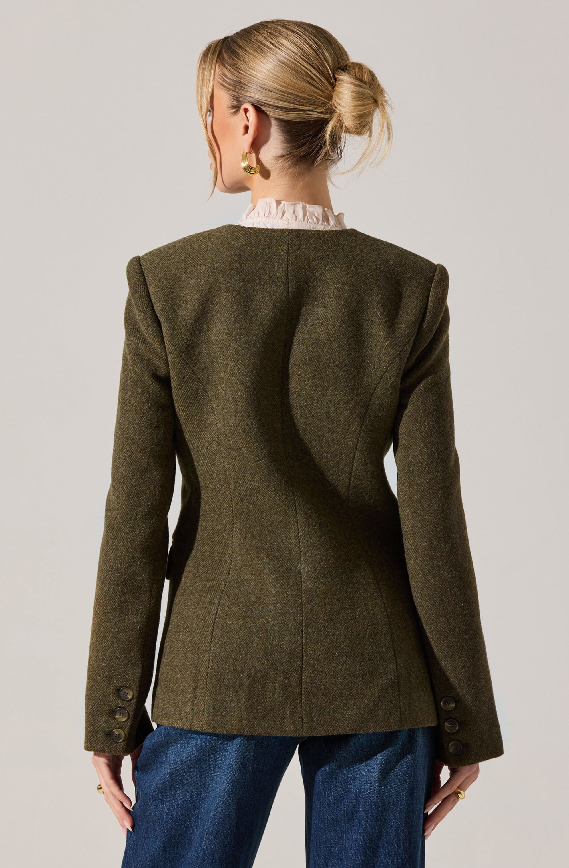 A woman is wearing a dark green, tailored blazer with structured, padded shoulders and detailed sleeve buttons. Underneath, she has a light pink, ruffled collared shirt. She also has on blue jeans. She is accessorized with gold hoop earrings and rings, and her hair is styled in a low, messy bun.