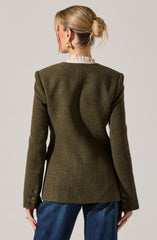 A woman is wearing a dark green, tailored blazer with structured, padded shoulders and detailed sleeve buttons. Underneath, she has a light pink, ruffled collared shirt. She also has on blue jeans. She is accessorized with gold hoop earrings and rings, and her hair is styled in a low, messy bun.