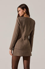 A woman is wearing a fitted brown blazer with a small checkered pattern, featuring structured shoulders and button details on the cuffs. She pairs the blazer with black sheer tights. She accessorizes with large gold earrings and a chunky gold chain necklace.