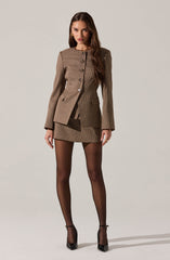 A woman is wearing a tailored, brown checkered blazer with a structured fit, featuring a row of buttons down the front and flap pockets at the hips. She pairs it with a matching mini skirt, sheer black tights, and black high-heeled shoes. The outfit is accessorized with gold earrings and a necklace.