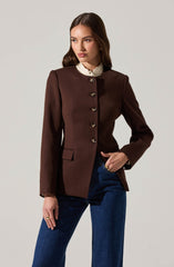 A woman is wearing a fitted, dark brown blazer with structured shoulders and a high neckline. The blazer features a row of large, shiny buttons down the front and has a small pocket flap on the left side. Underneath, she has on a white, ruffled collared blouse. She pairs the blazer with dark blue, straight-leg jeans.