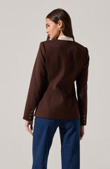 A woman is wearing a tailored dark brown blazer with structured seams and button detail on the cuffs. The blazer features a white collar peeking out from underneath. She is also wearing blue jeans.