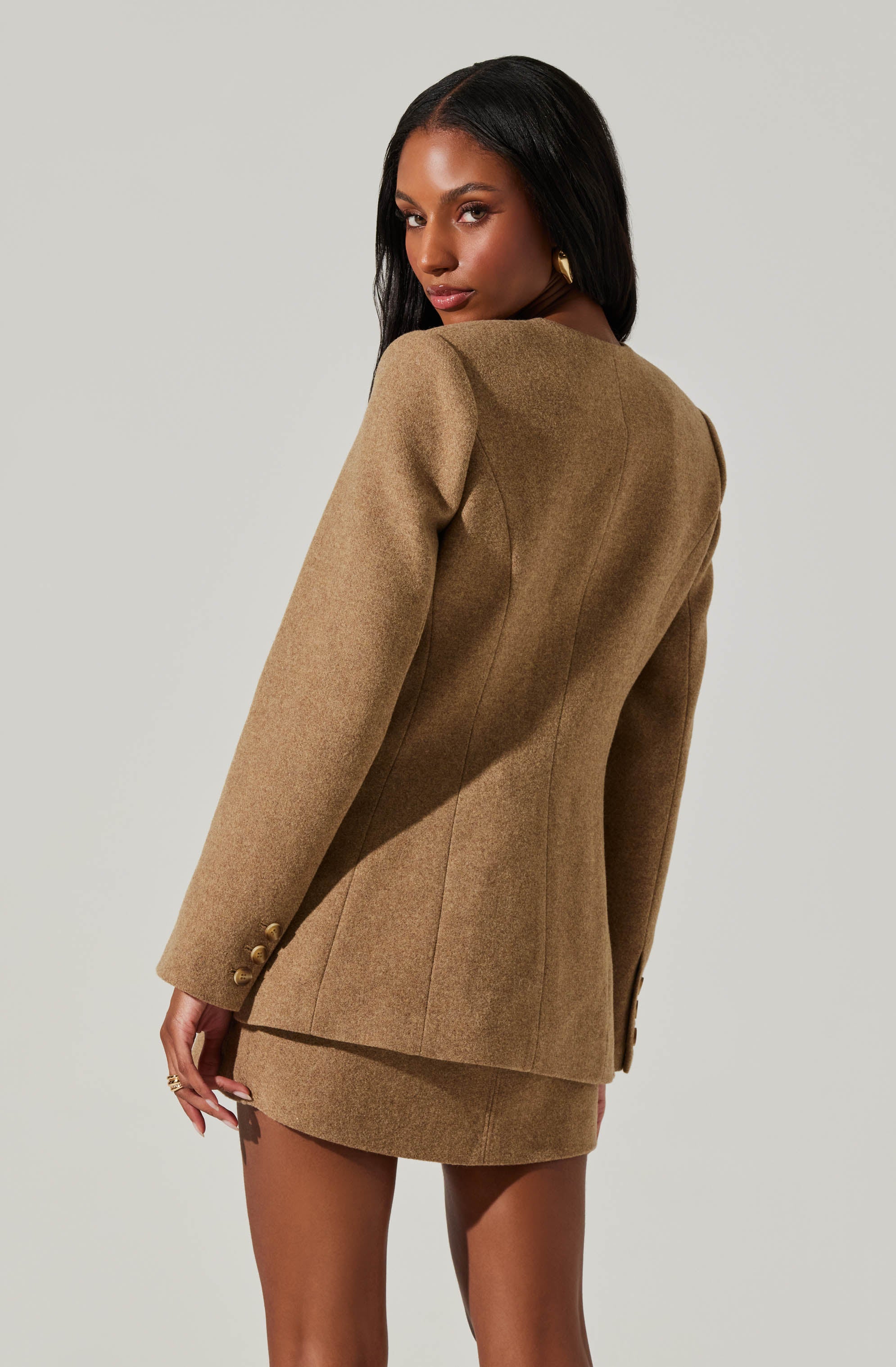 A woman is wearing a tan-colored, tailored blazer with a matching mini skirt. The blazer features a structured fit, with seam details and gold buttons on the sleeves. The outfit appears to be made of a wool or wool-blend fabric.