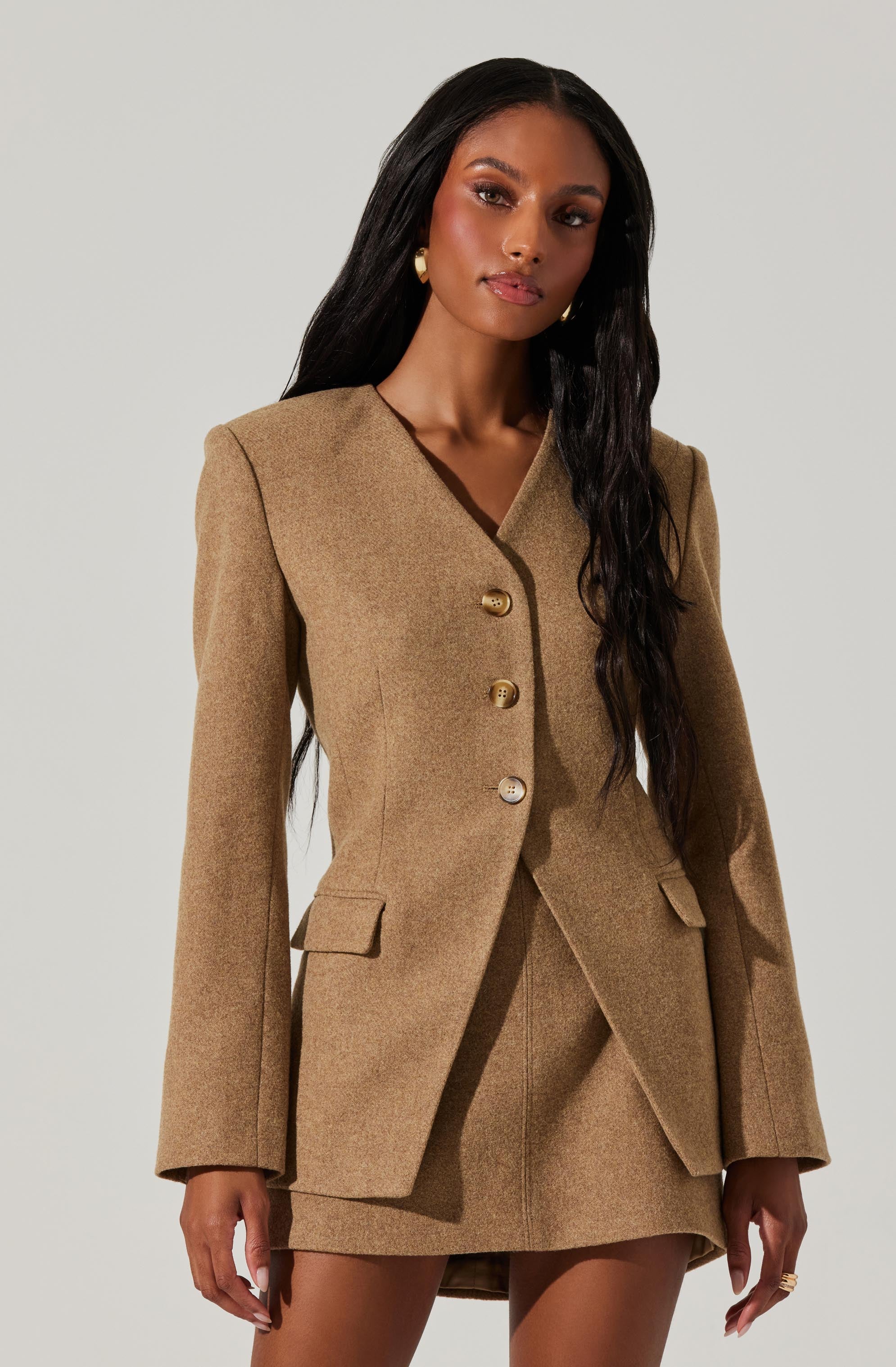 A woman is wearing a tan, tailored blazer with padded shoulders and a notched lapel. The blazer features three large, round buttons and two flap pockets at the front. She is also wearing a matching tan skirt that is slightly longer in the back, creating a high-low effect. The outfit is sophisticated and chic.