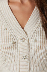 A close-up view of a cream-colored, knitted cardigan featuring pearl and rhinestone embellishments. The cardigan has a ribbed collar and vertical ribbing throughout the fabric. The decorative embellishments include pearl accents and floral-shaped embellishments with pearls and rhinestones.