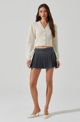 A woman is wearing an ivory-colored, long-sleeved, V-neck cardigan with ribbed knit texture and pearl-like button details down the front. She is also wearing a high-waisted, dark gray pleated mini skirt with a belt loop and buckle detail. She completes the outfit with white pointed-toe high heels.