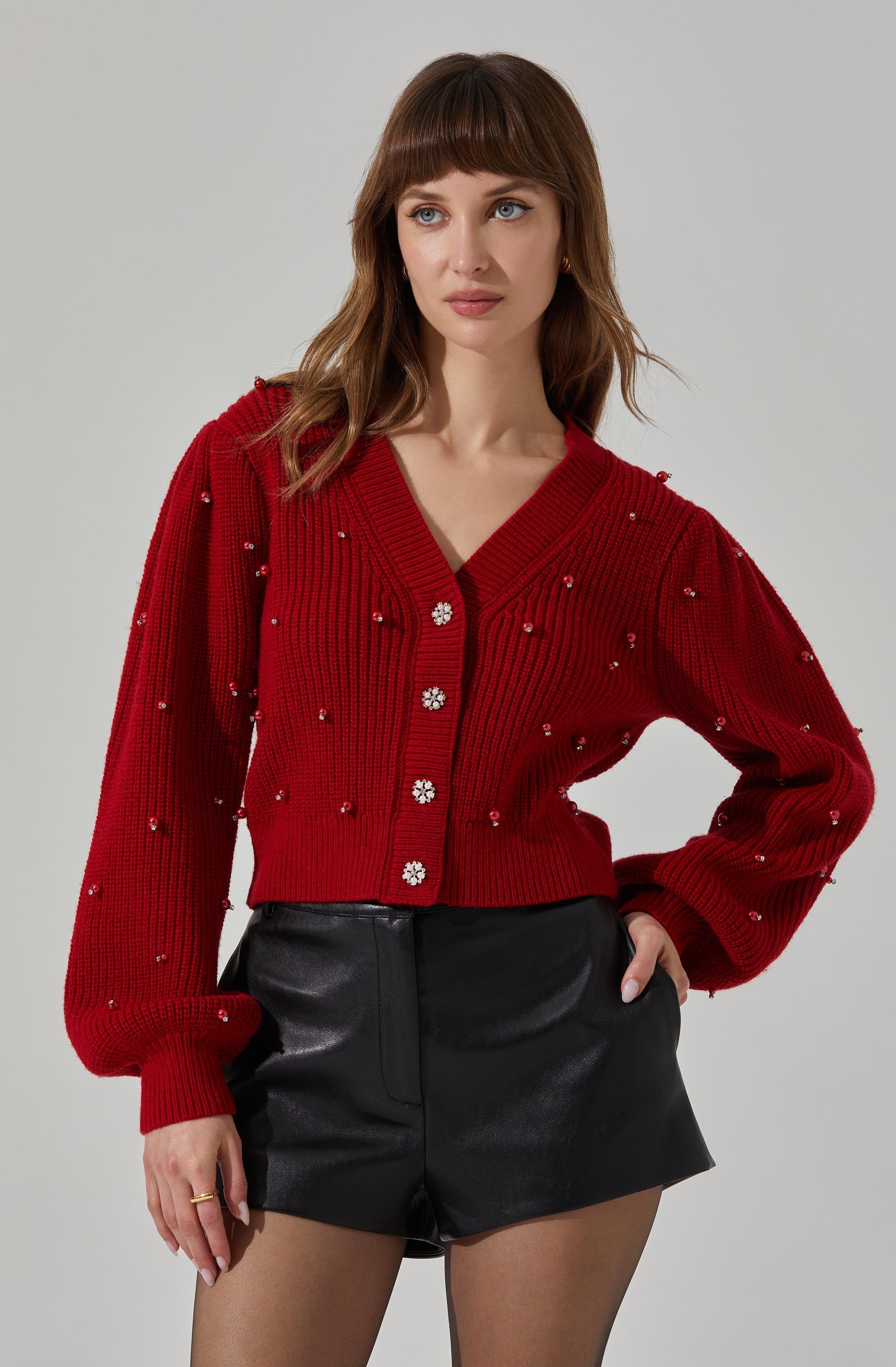 A woman is wearing a red, knitted cardigan with large floral-shaped buttons and small decorative beads scattered across the fabric. The cardigan has a V-neckline, long puffed sleeves, and ribbed cuffs and hem. She is paired it with black leather shorts.