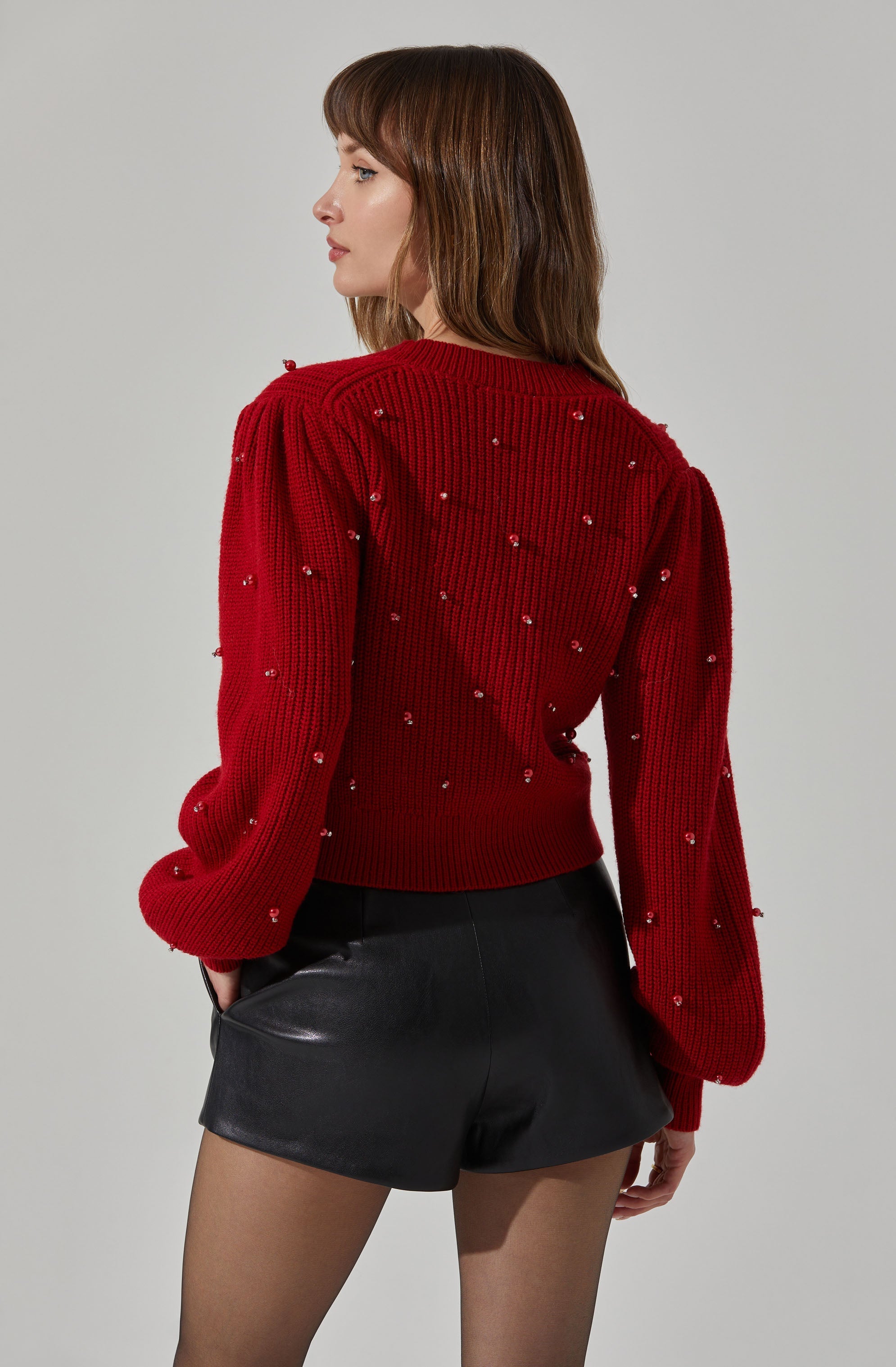 A woman is wearing a red knit sweater decorated with small pearl embellishments and black leather shorts. The sweater has a ribbed texture with slightly puffed long sleeves. She is also wearing sheer black tights.