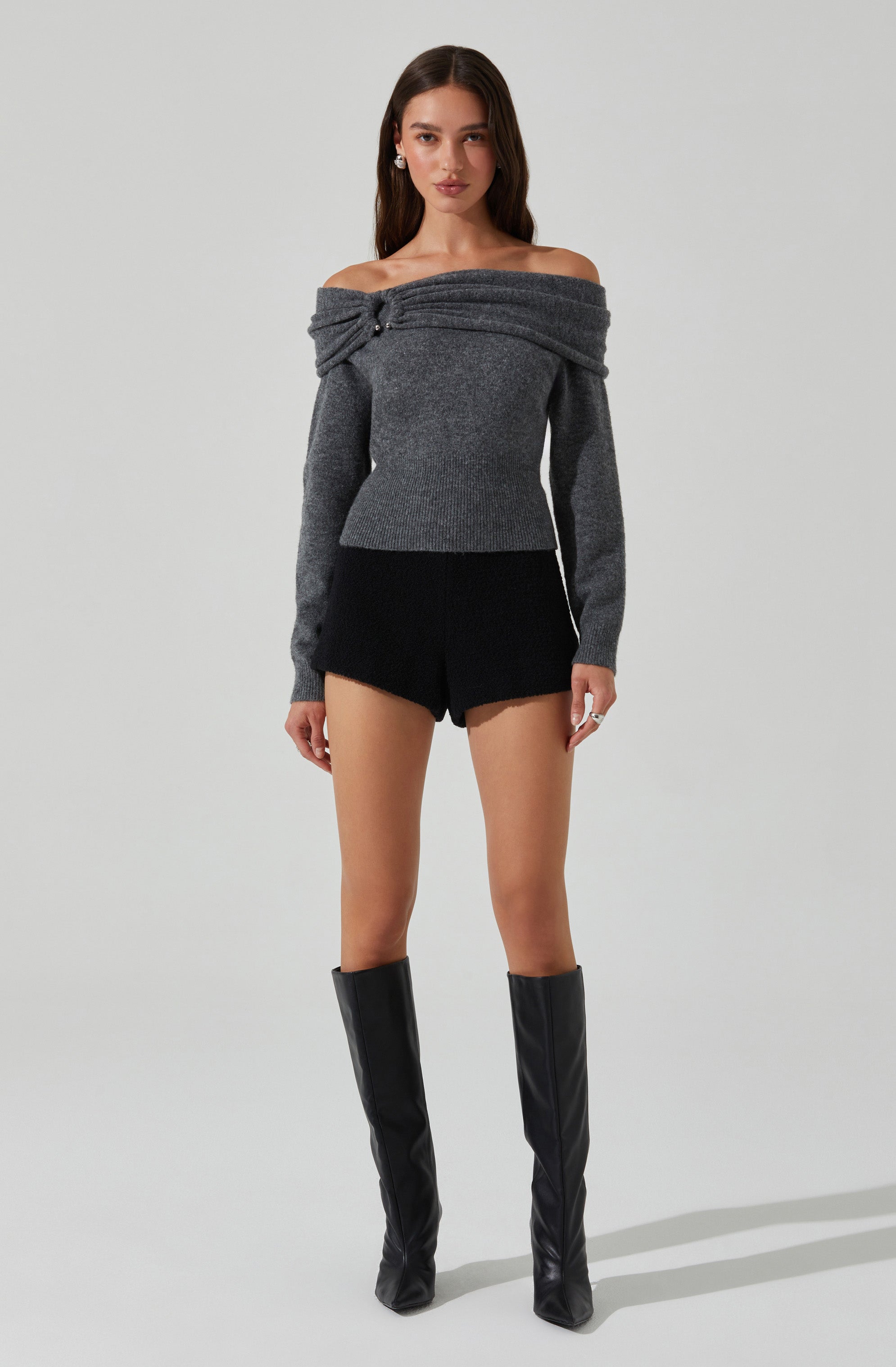 A woman is wearing an off-the-shoulder, long-sleeved gray sweater with a knot detail at the shoulder. She pairs it with black shorts and tall, black knee-high boots.
