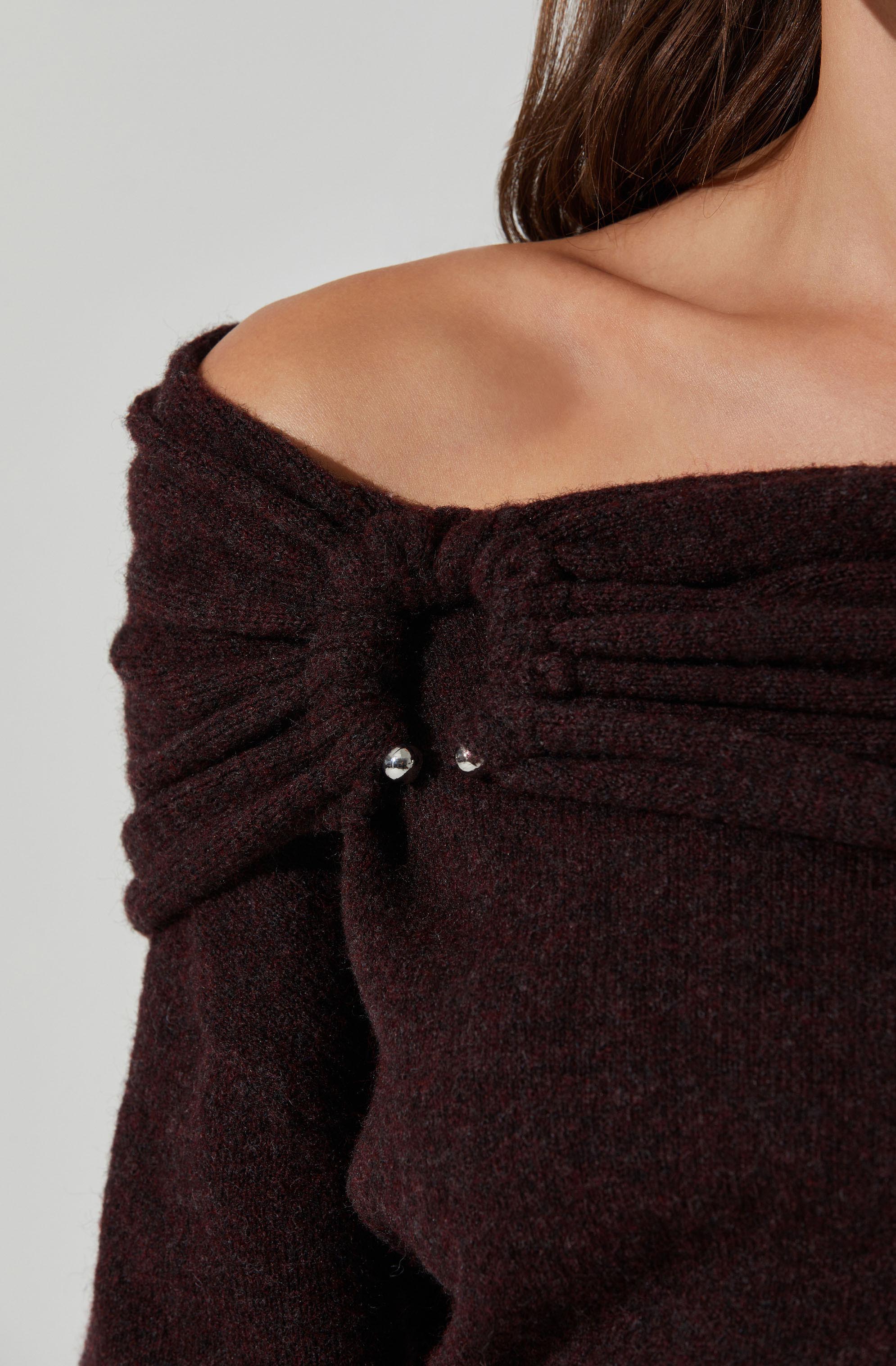 A person is wearing a dark brown, off-the-shoulder sweater with textured, ribbed detailing along the top edge. The sweater has gathered fabric at the shoulder with small decorative metallic beads.