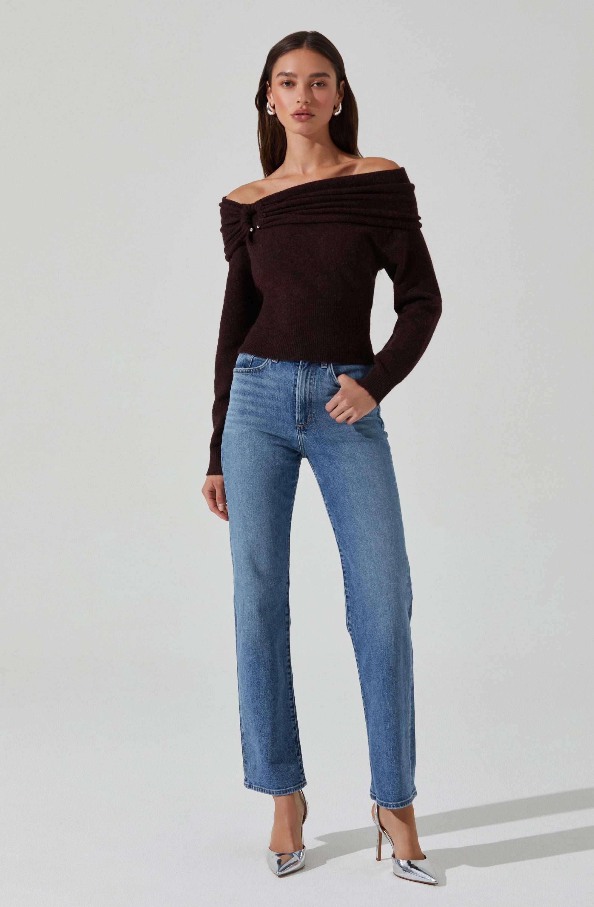 A woman is wearing an off-the-shoulder, long-sleeved, dark brown sweater with a textured, ribbed design across the top. She pairs it with high-waisted, light blue jeans and silver, pointed-toe high heels. Her dark brown hair is styled straight, and she accessorizes with hoop earrings.