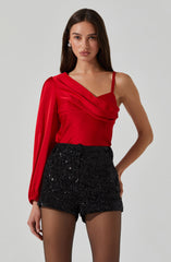 A woman is wearing a red satin top with a draped, asymmetrical design featuring a single shoulder strap on her right side and a long sleeve on her left arm. She pairs it with high-waisted black sequined shorts.
