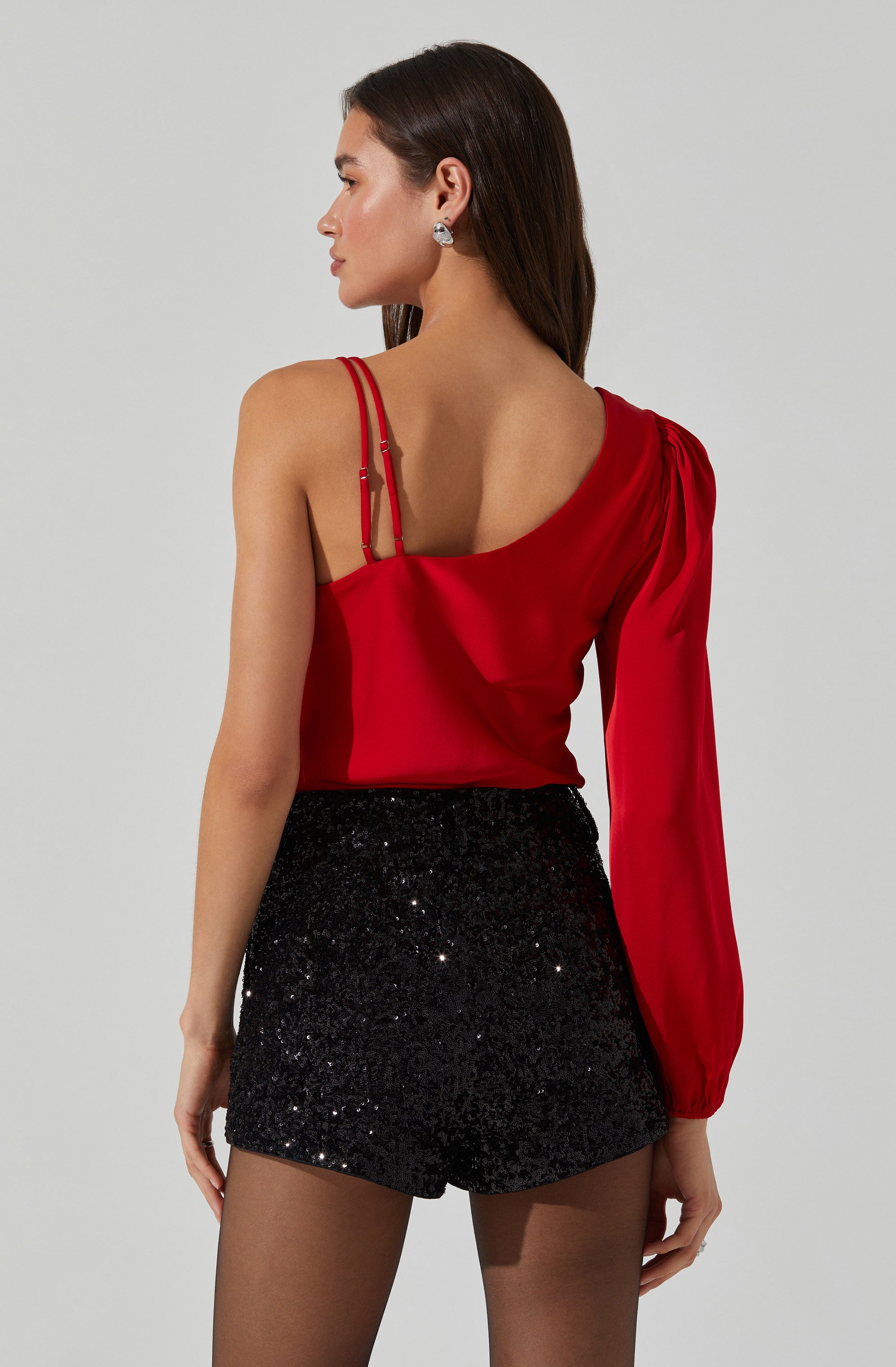 A woman is wearing a red satin top with spaghetti straps on the left shoulder and a long, gathered sleeve on the right arm. She pairs it with high-waisted black shorts that are covered in black sequins, creating a shiny, glittery appearance.