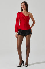 A woman is wearing a red top with asymmetrical design, featuring a draped, one-shoulder style with one long sleeve and the other shoulder strap. She pairs this with black, high-waisted shorts decorated with sequins, and completes the look with black high heels that have pointed toes and decorative details on the front.