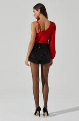 A woman is wearing a red, one-shoulder top with a single long sleeve and double spaghetti straps on the other shoulder. She pairs it with black high-waisted shorts that are covered in sparkly sequins. She completes her outfit with sheer black tights and black high-heeled shoes.