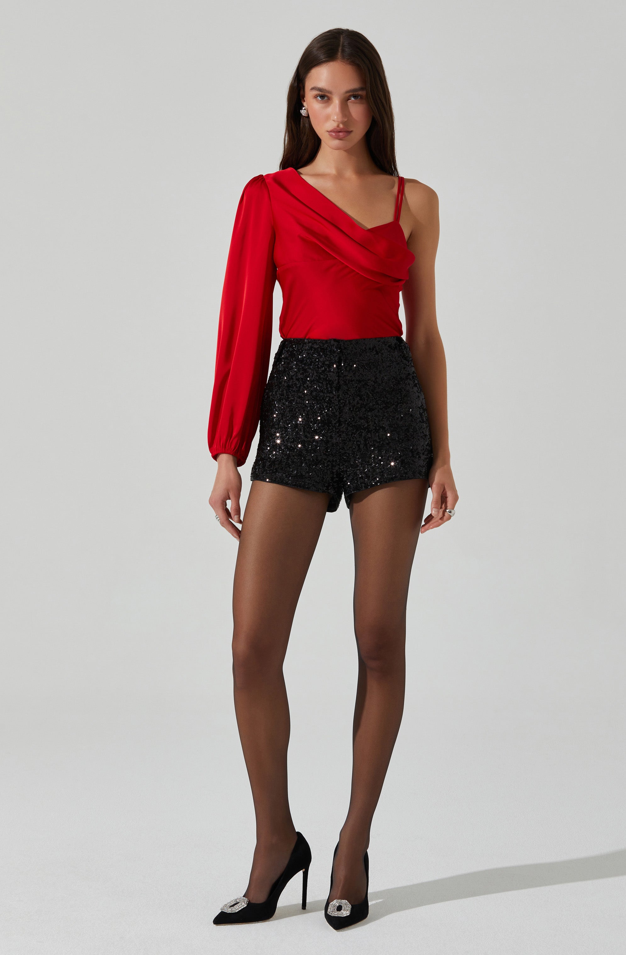 A woman is wearing a red, one-shoulder top with a draped design and long balloon sleeve on one side. She pairs it with high-waisted black sequined shorts. She completes the outfit with black high heels that have decorative embellishments on the toe.