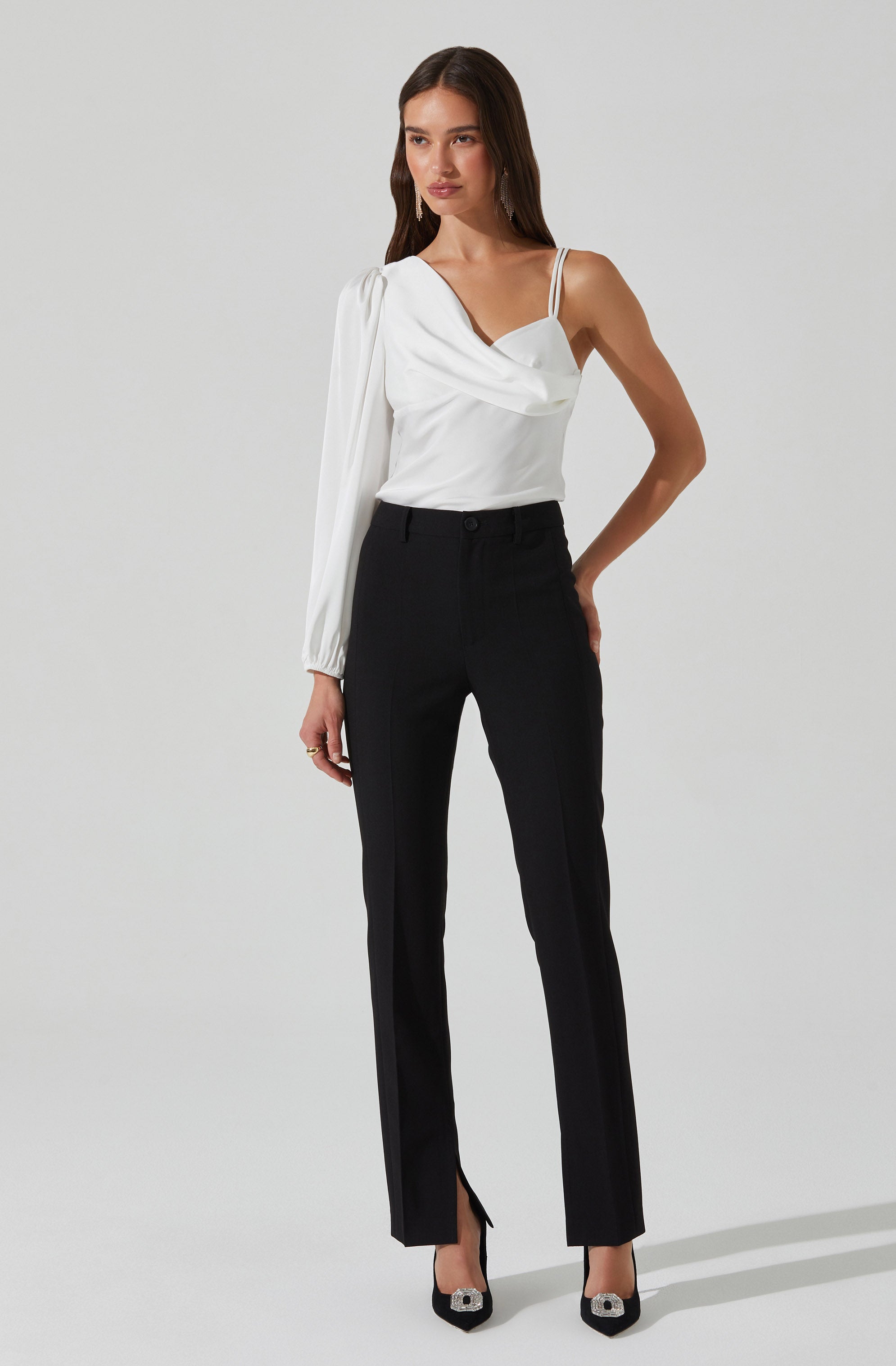 A woman is wearing a white blouse with a draped neckline and spaghetti straps, paired with high-waisted black trousers that have a front slit at the hem. She is also wearing black high-heeled shoes with decorative embellishments.